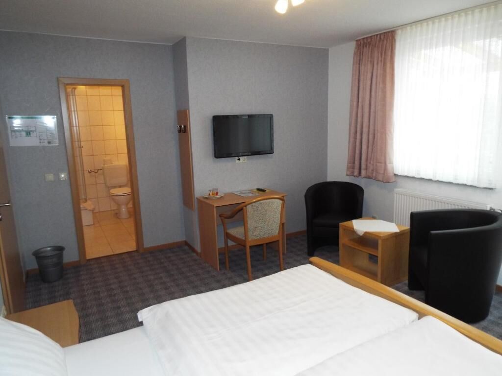 A hotel room with a bed, a desk with a chair, a television, and a bathroom visible through an open door.
