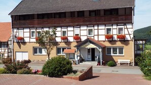 Un edificio de dos pisos con fachada tradicional de entramado de madera, una entrada central con un pequeño porche y macetas con flores en los alféizares de las ventanas.