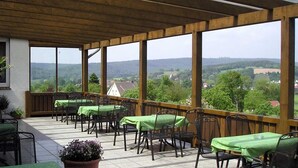 Un espacio comedor al aire libre techado, con mesas y sillas, techo de madera y vista a un paisaje rural.