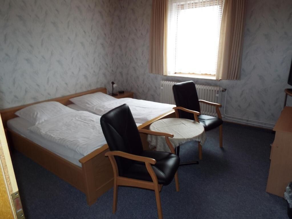 A room with two single beds, a round table, two chairs, a radiator, and a window with curtains.
