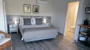 Un dormitorio con cama, mesitas de noche, un taburete, una silla de mimbre, piso de madera, baño con inodoro y repisa con utensilios de cocina.