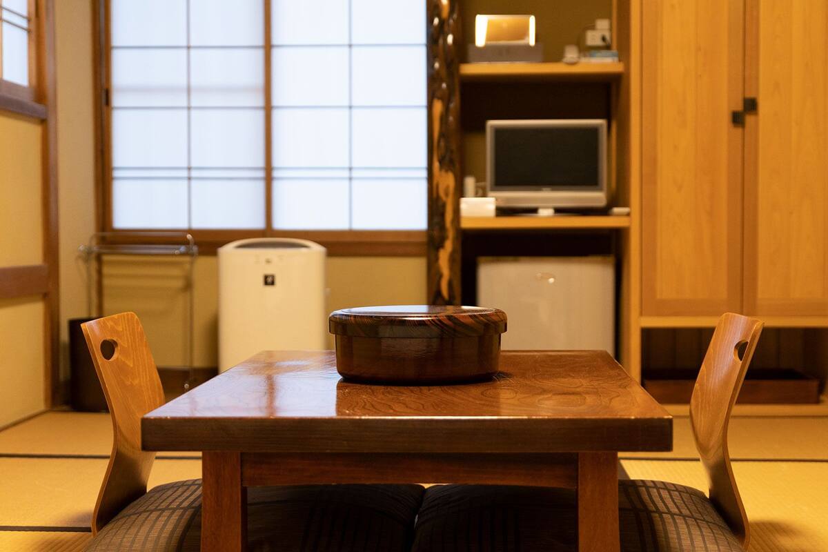 A traditional Japanese room with tatami mat flooring, a wooden table with a dark wooden bowl, and a television on a shelf.