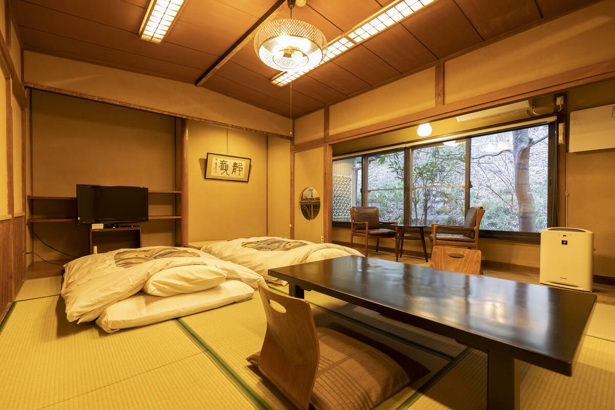 A traditional Japanese room with tatami flooring, a low table, a TV, and a window with a view of trees.