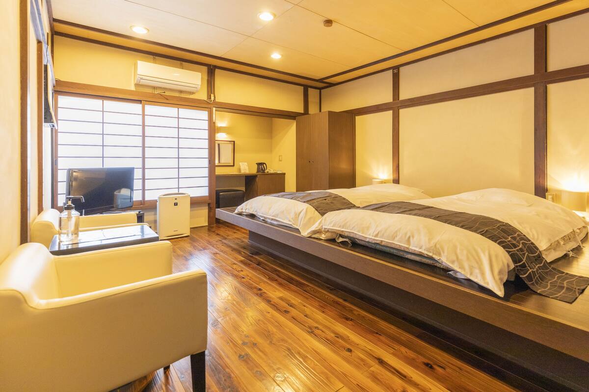 A hotel room with a wooden floor, a bed with white and patterned bedding, a desk with a chair, and a television.