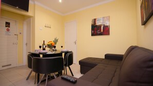 Uma sala de estar com um sofá marrom, uma mesa de jantar com cadeiras e uma TV fixada na parede.