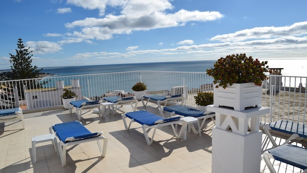 Uma área de estar ao ar livre ensolarada com espreguiçadeiras brancas e azuis, um vaso branco com arbusto e vista para o mar.