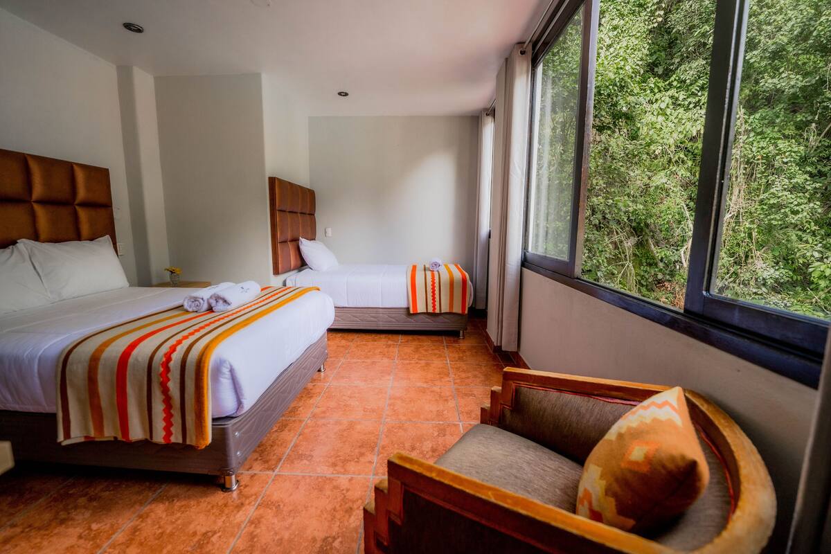 Quarto de hotel com duas camas, uma janela ampla com vista para áreas verdes e uma cadeira de madeira com almofada.