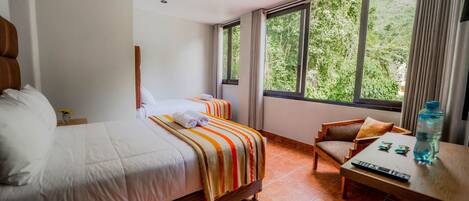 A hotel room with two beds, a wooden chair, a small table, and a window with a view of greenery.