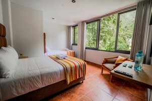 Habitación de hotel con dos camas, una silla de madera, una mesita y una ventana con vistas a zonas verdes.
