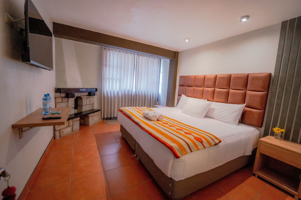 Quarto de hotel com cama, televisão na parede, uma mesinha com uma garrafa e janela com cortinas.