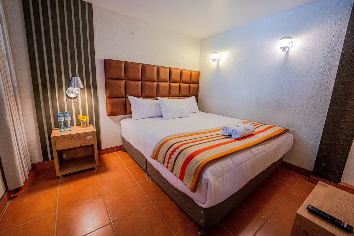 Cama bem arrumada com manta listrada, mesa de cabeceira de madeira com abajur e controle remoto sobre uma pequena mesa.