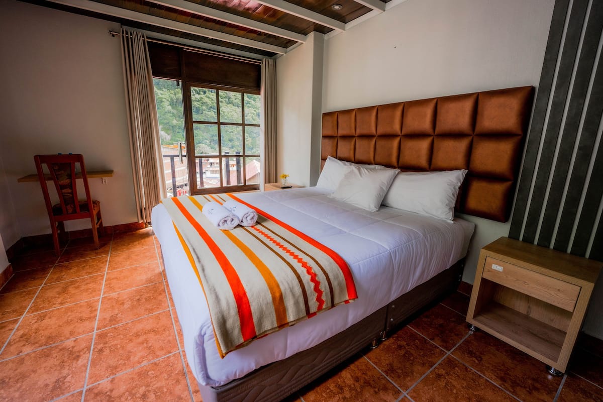 Quarto de hotel com uma cama grande, uma mesinha de cabeceira de madeira, uma cadeira e uma janela com cortinas.