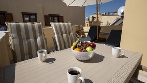 Un tavolo con due sedie a righe, una ciotola di frutta e due tazze bianche su una terrazza illuminata dal sole con un ombrellone.