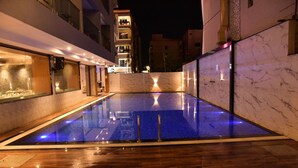 A swimming pool with a wooden deck, surrounded by walls with marble patterns, and a building with balconies in the background. - SWARNA HOTEL & RESORT (Kanthi)