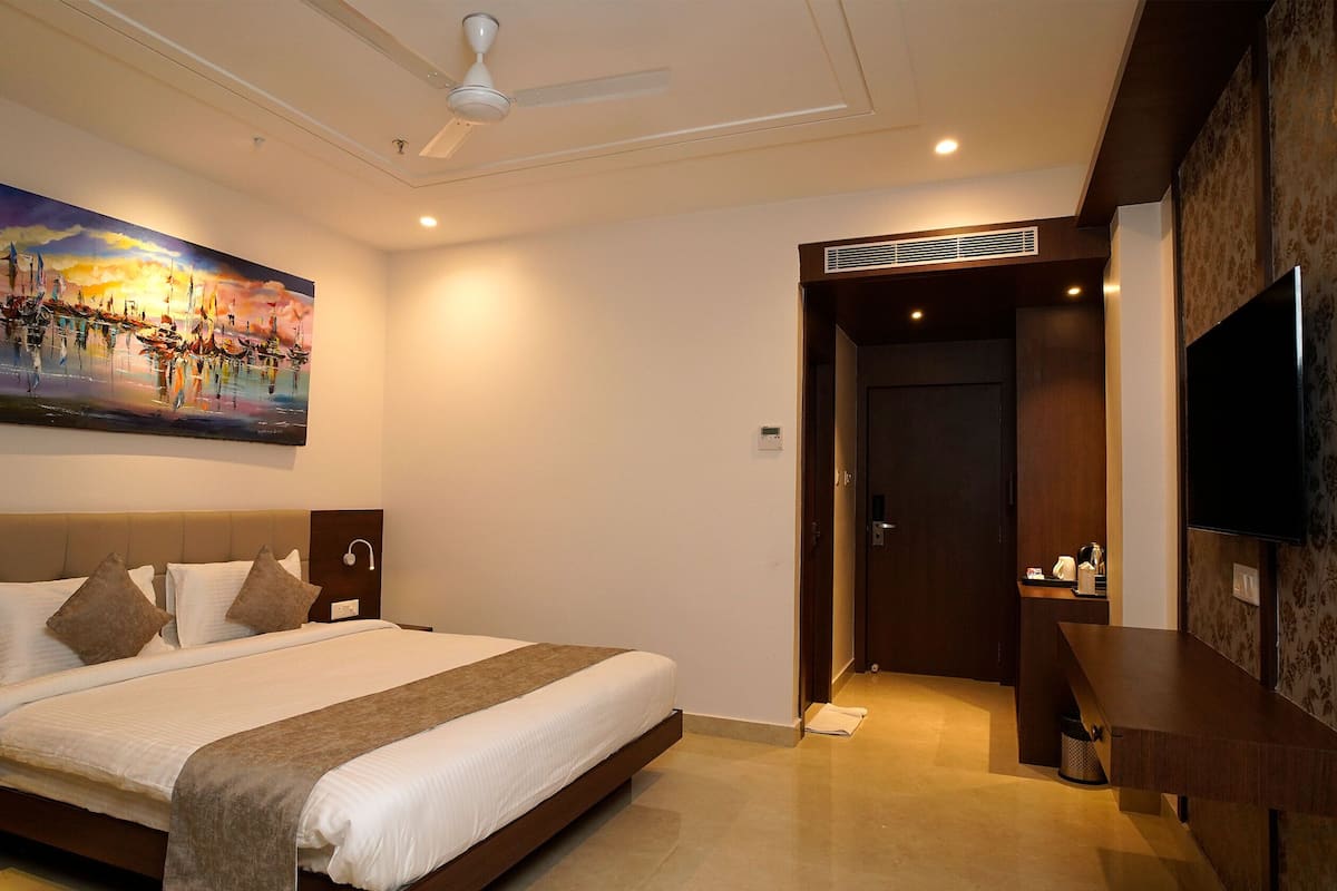 A hotel room with a large bed, a painting on the wall, a ceiling fan, and a TV mounted on the wall.