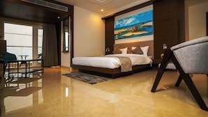 A hotel room with a bed, a desk, a chair, and a painting of a boat on a beach. - SWARNA HOTEL & RESORT (Kanthi)