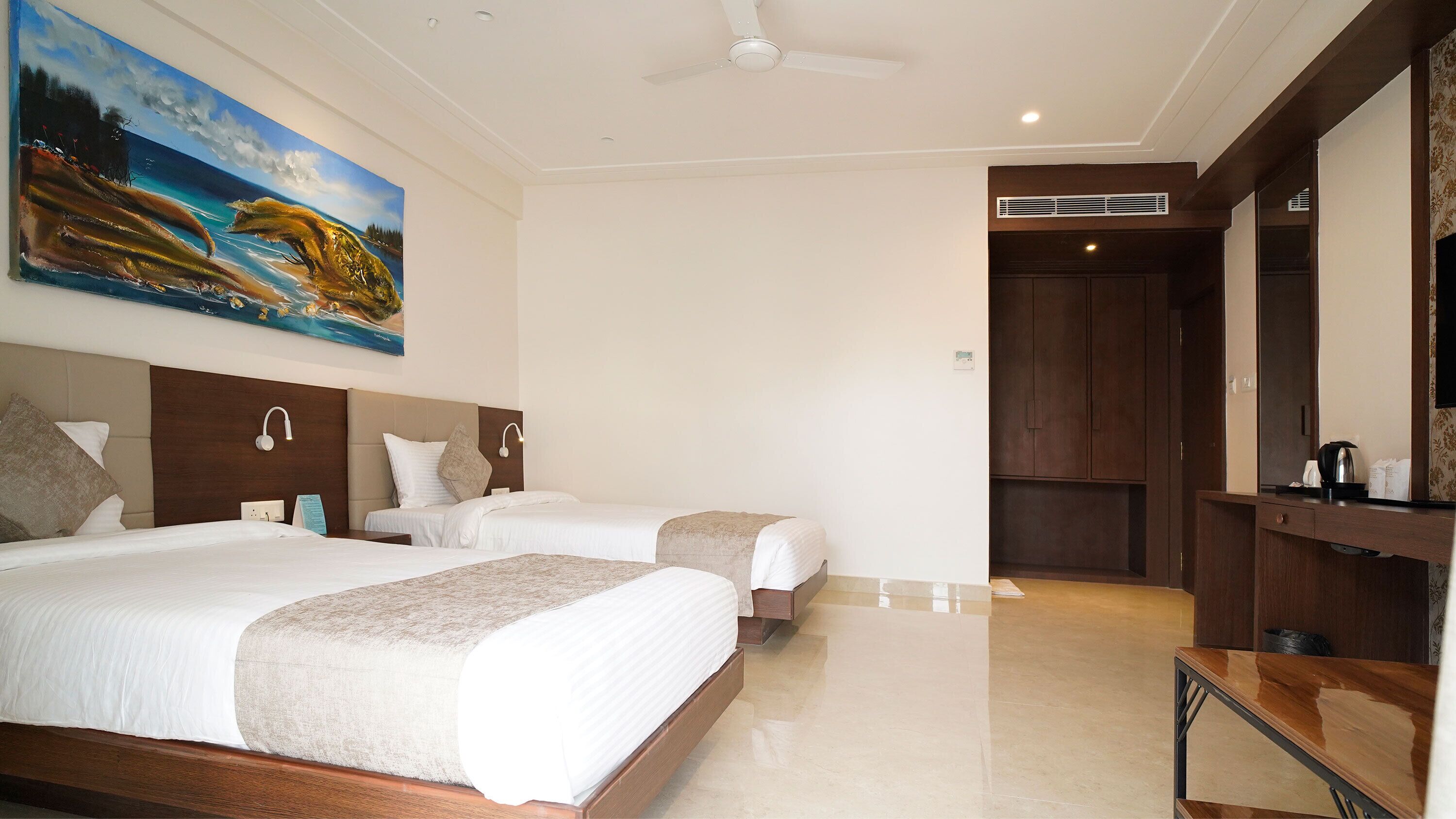 A hotel room with two beds, a painting of a landscape, a ceiling fan, and a wooden wardrobe.