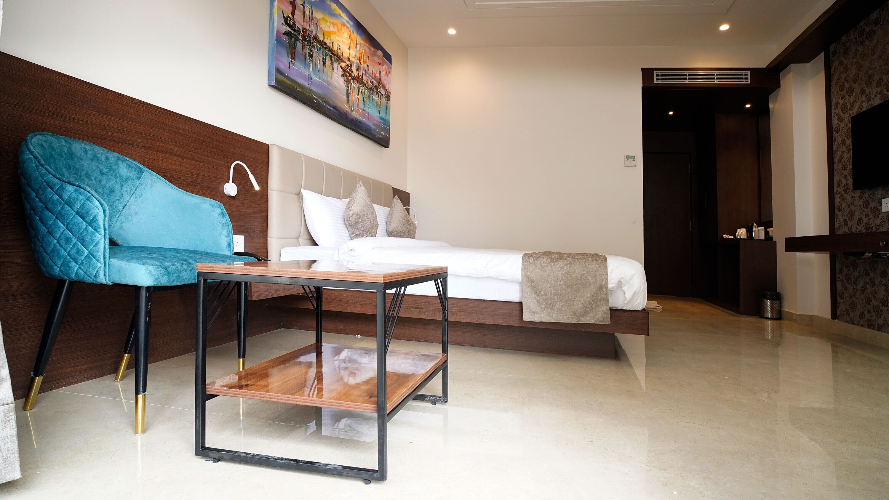 A modern hotel room with a bed, a blue armchair, a wooden table, and a painting on the wall.
