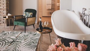 Un baño moderno con una bañera independiente, un sillón verde, una mesita con una botella y vasos, y una alfombra estampada con motivos florales.