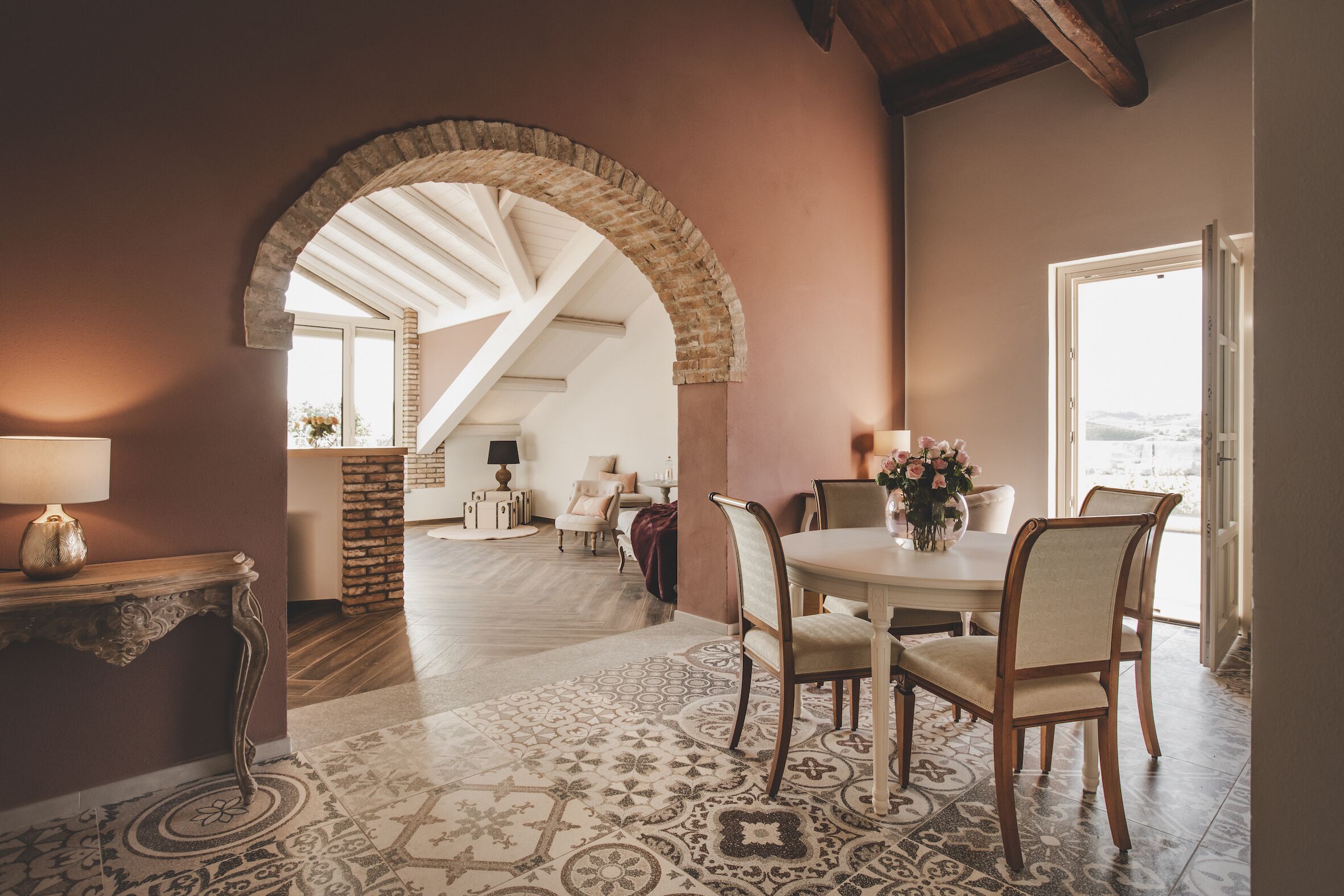 A dining area with a round table, chairs, and a patterned floor. An arched doorway leads to another room.