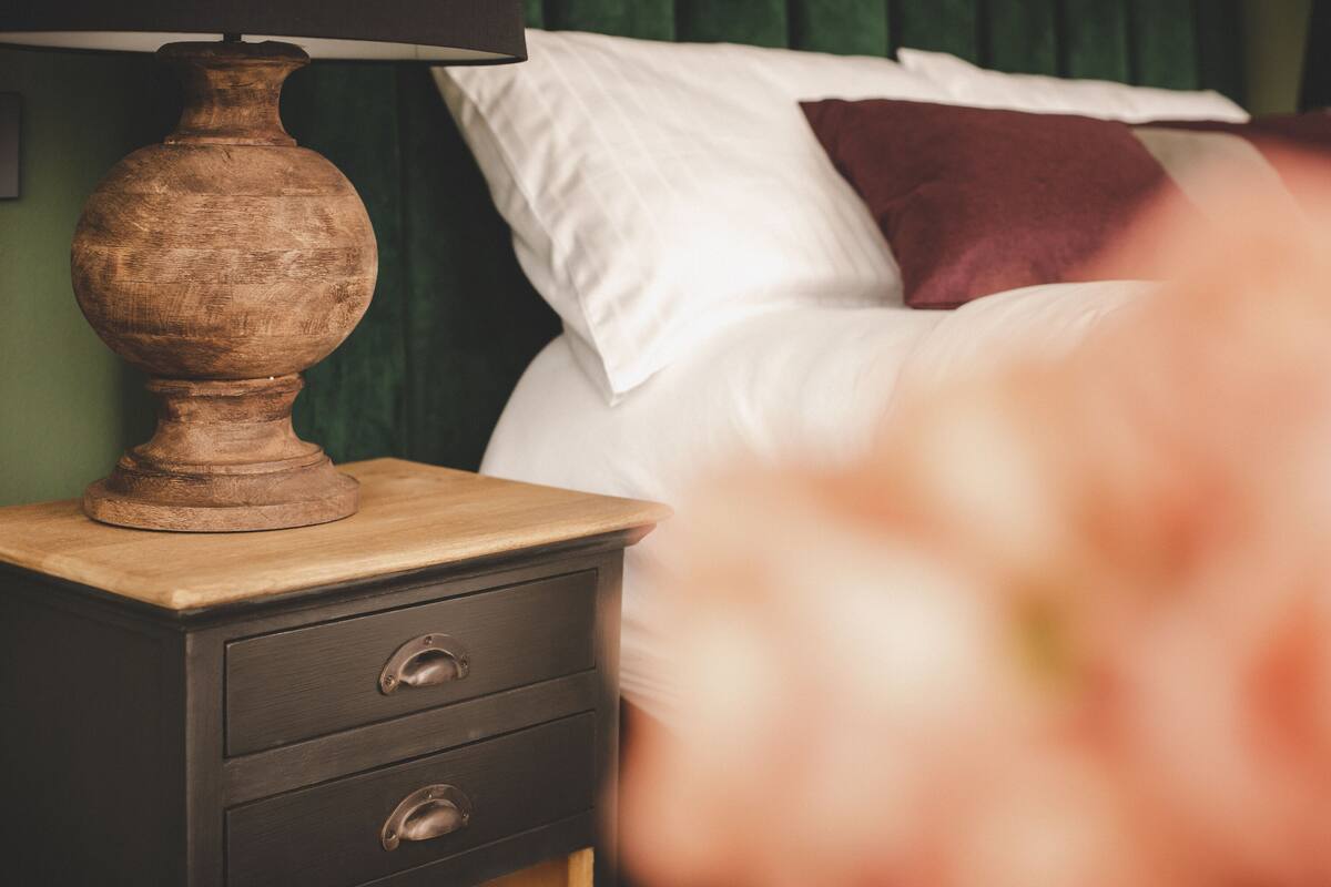 A wooden bedside table with a lamp, a bed with white and red pillows, and a green curtain in the background.