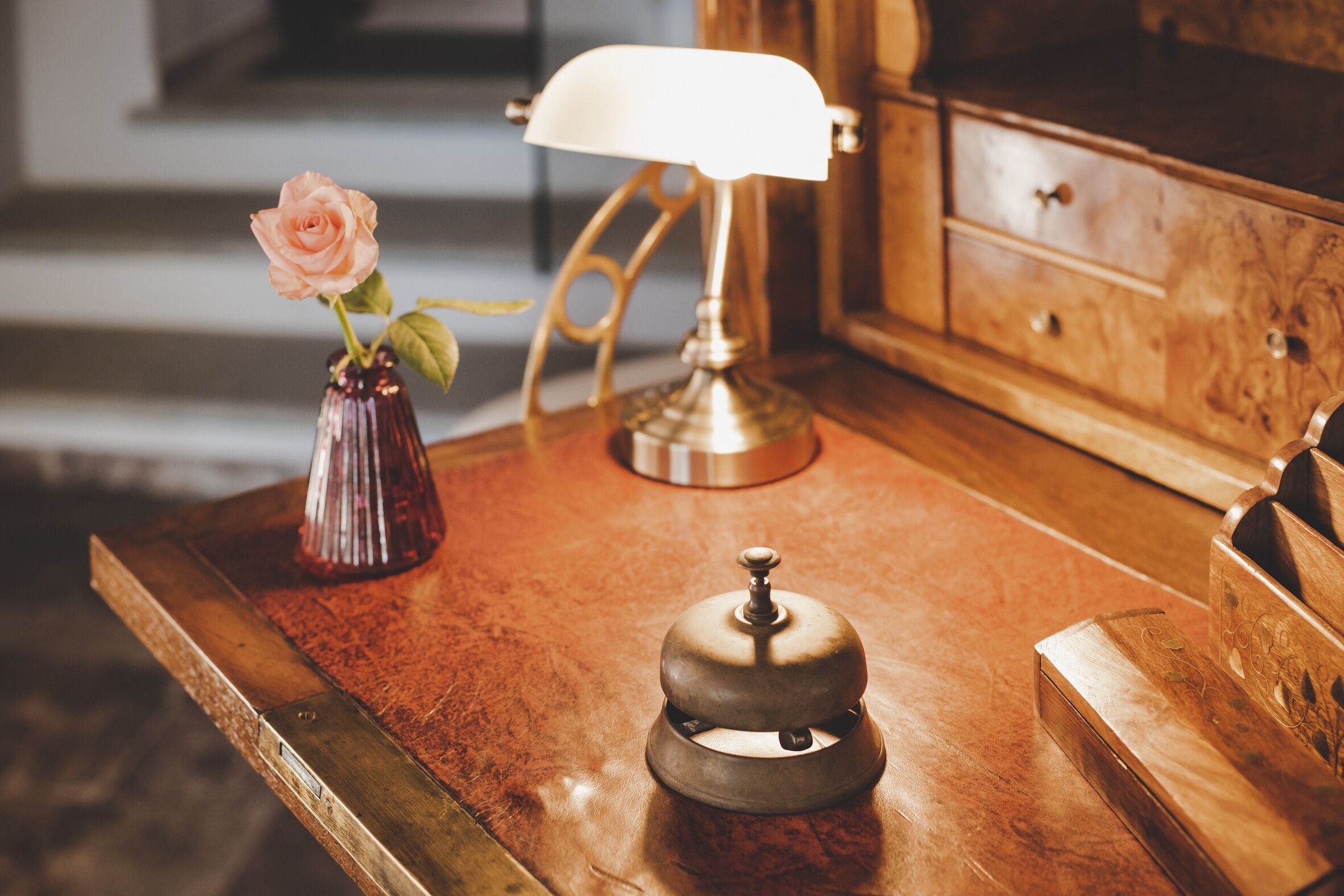 Un escritorio de madera con un teléfono de bronce, un florero con una rosa y una lámpara.