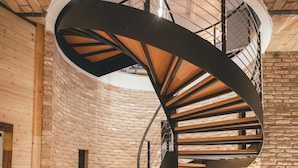 Una escalera de caracol con escalones de madera y barandillas metálicas en una habitación con paredes de ladrillo visto y suelo de baldosas.