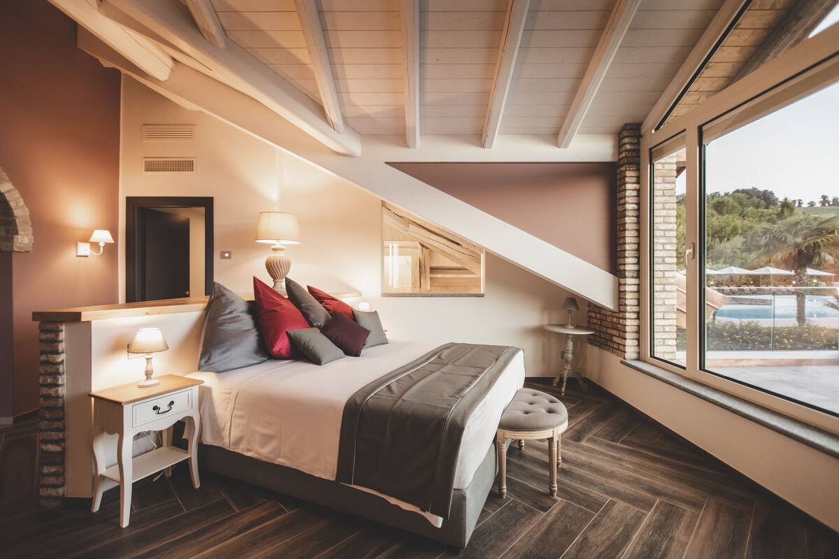 A bedroom with a bed, bedside tables, a lamp, a bench, and a view of the outdoors.