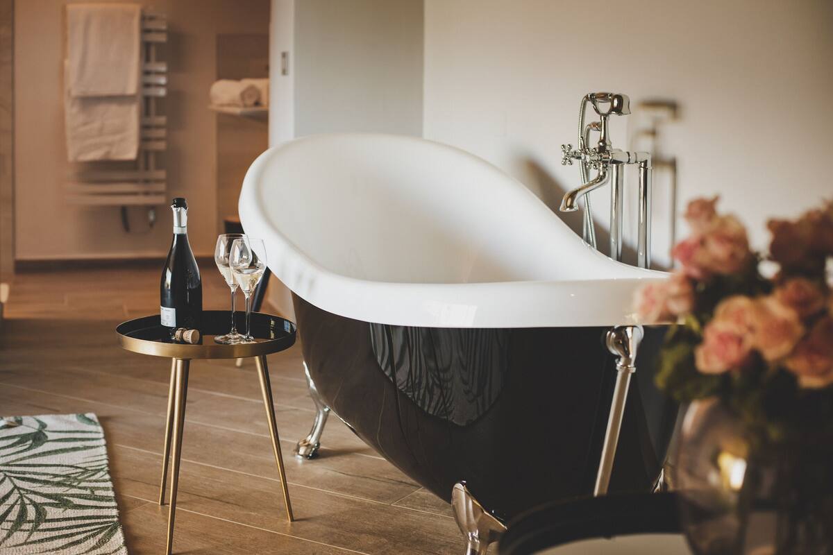 A modern bathroom with a freestanding bathtub, a small round table with a bottle and glasses, and a vase of flowers.