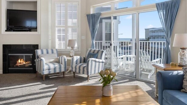 Un salon avec une cheminée, des fauteuils à rayures, un canapé, un téléviseur et un balcon avec des chaises blanches.