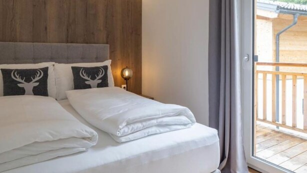 Una cama bien tendida con sábanas blancas y dos almohadas decorativas, una lámpara de noche y vistas a una terraza de madera a través de un ventanal grande.