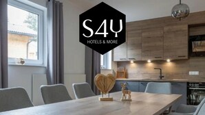Un comedor moderno con mesa de madera y sillas tapizadas en gris, una cocina al fondo, y el logotipo de S4U Hotels & More.