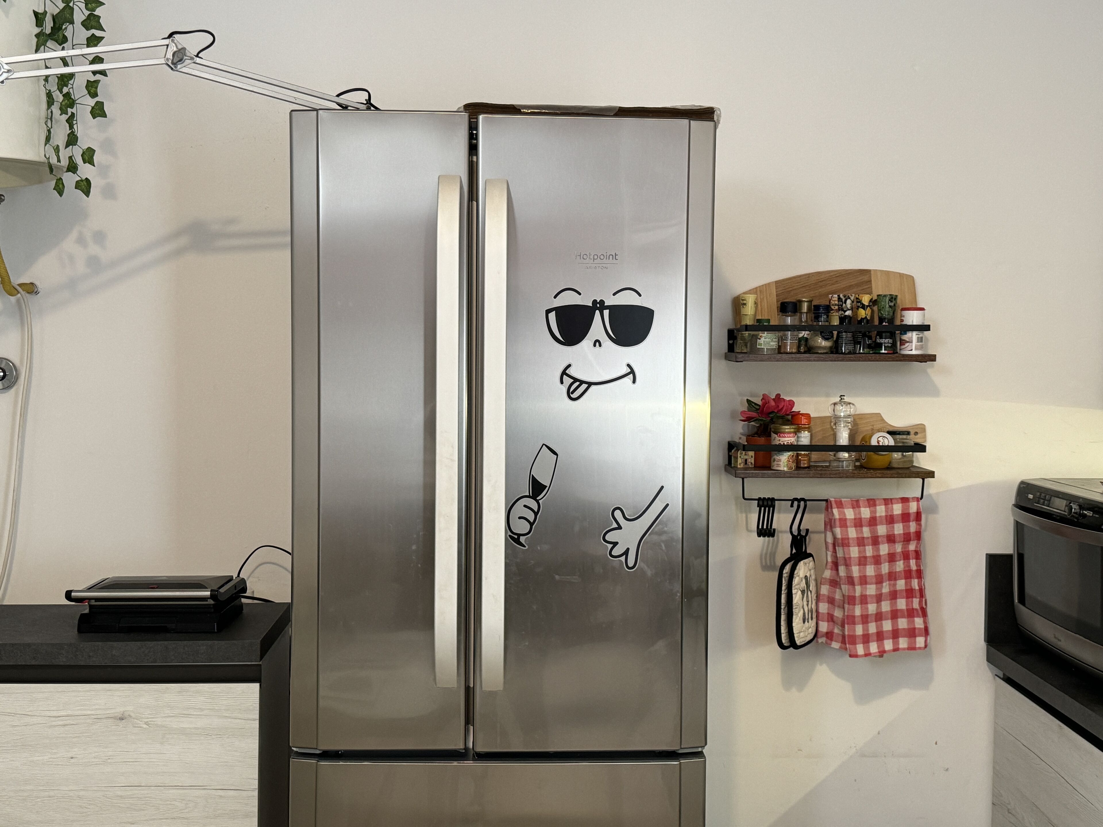A stainless steel refrigerator with a cartoon face sticker, a kitchen counter, a microwave, and a small shelf with kitchen items.