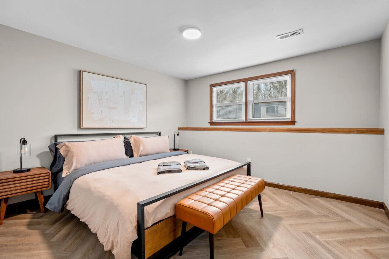 A bedroom with a bed, bench, bedside table, lamp, and window.