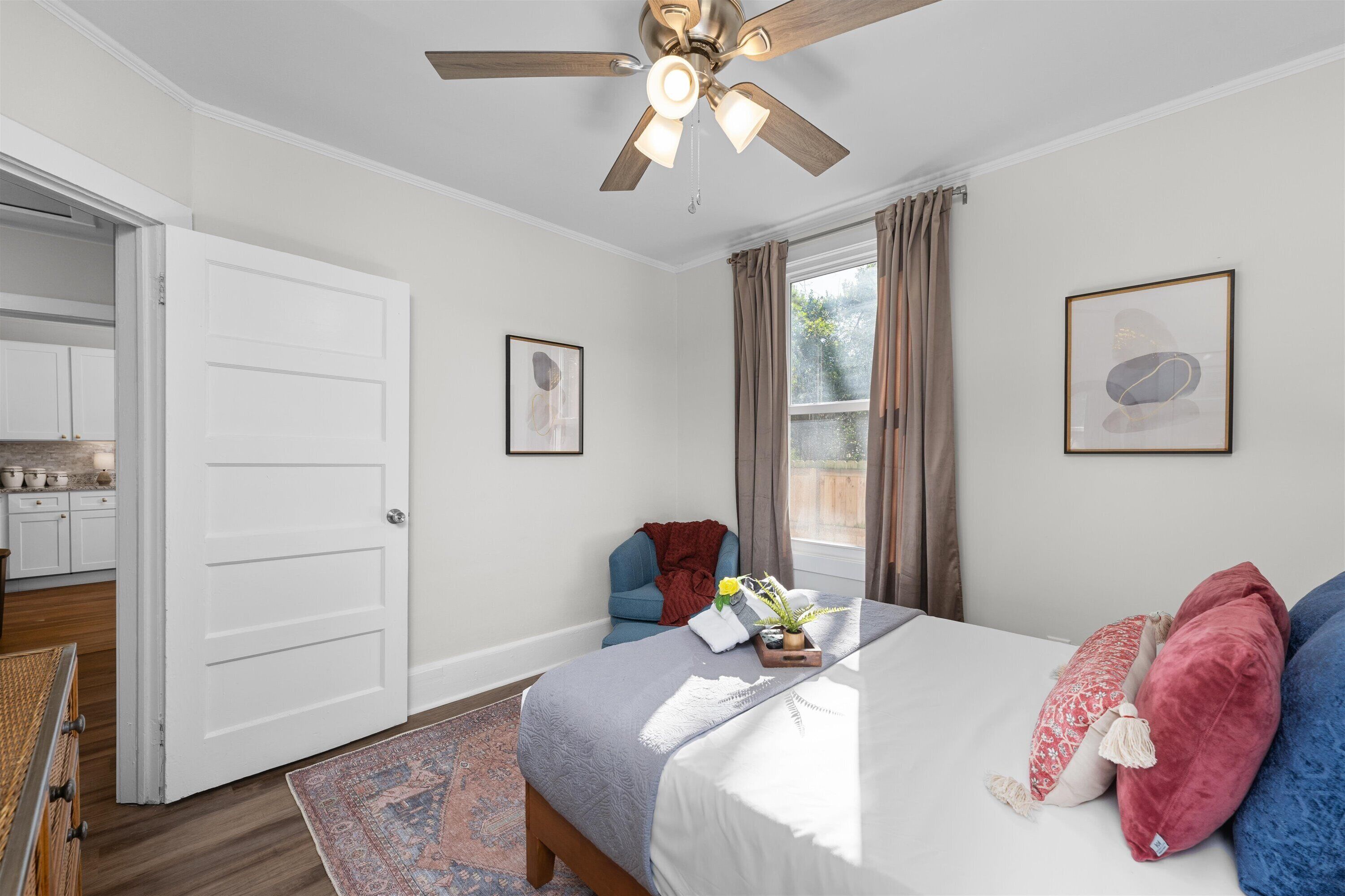 Ein Schlafzimmer mit einem Bett, einem Deckenventilator, einem Fenster mit Vorhängen, einem Sessel und einem kleinen Tisch mit einer Pflanze.