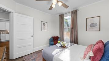 Ein Schlafzimmer mit einem Bett, einem Deckenventilator, einem Fenster mit Vorhängen, einem Sessel und einem kleinen Tisch mit einer Pflanze.