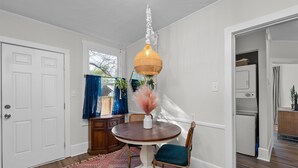 Uma área de refeições com uma mesa redonda de madeira, um vaso branco com flores secas e um candeeiro de ratã suspenso.