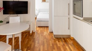 Ein kompaktes Hotelzimmer mit Holzboden, Flachbildfernseher, einem kleinen Esstisch, einem Bett und einer Küchenzeile mit Herd.