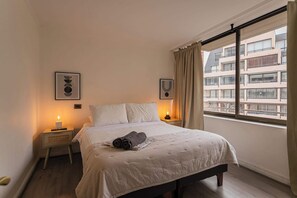 A bedroom with a bed, bedside lamps, a window with curtains, and a view of buildings. - El Golf: 2BR/2BA Family Apartment with Pool! (Las Condes)