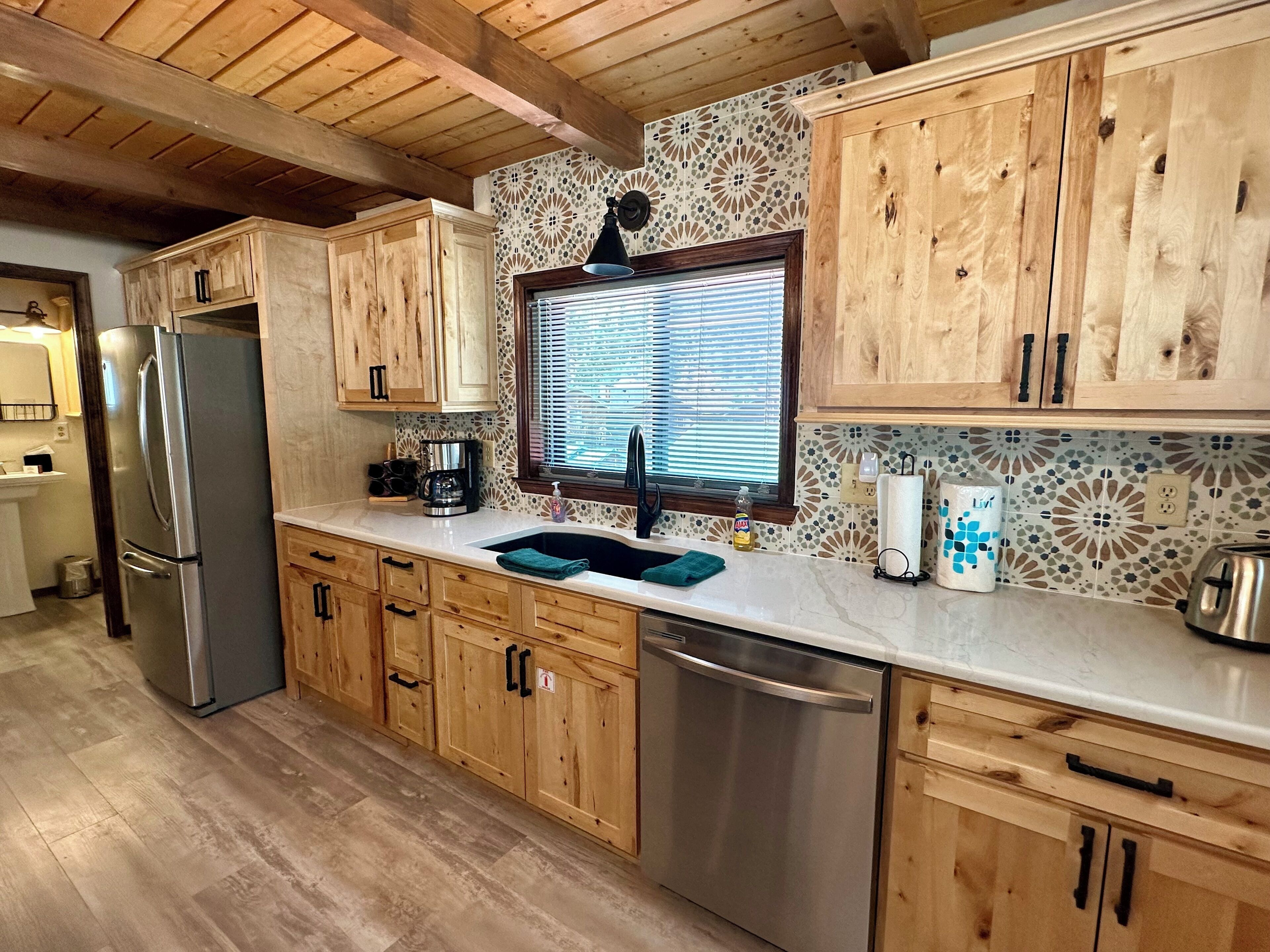 A kitchen with wooden cabinets, a stainless steel refrigerator, a dishwasher, and a window with blinds.