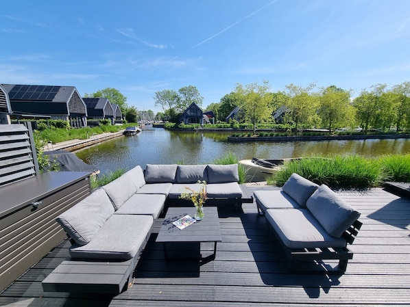 A deck with a sofa and a table, overlooking a canal with boats and houses.