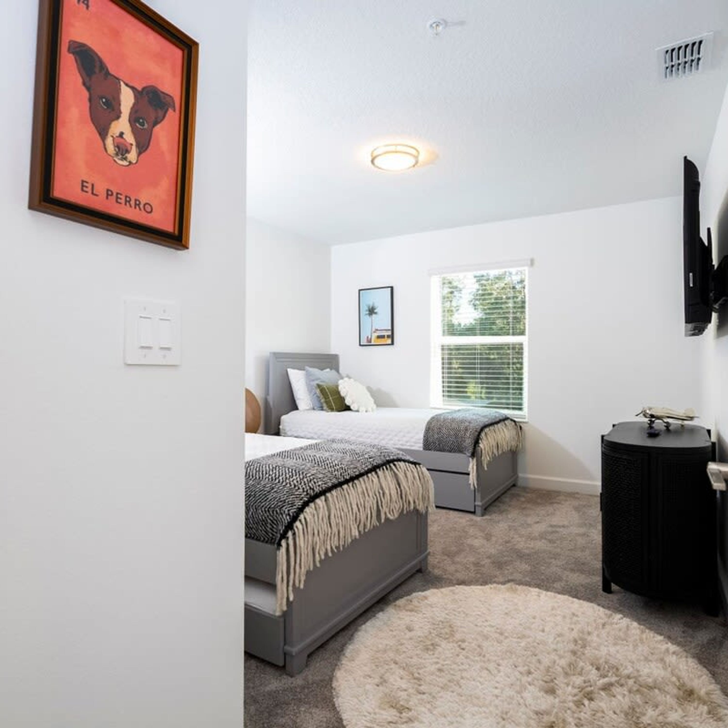 A bedroom with two beds, a large window, a ceiling light, and a framed picture on the wall.