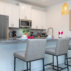 A modern kitchen with white cabinets, a stainless steel refrigerator, a microwave, and a large island with bar stools. - Gorgeous 4 Bd w Pool Storey Lake Resort 2485 (Kissimmee)