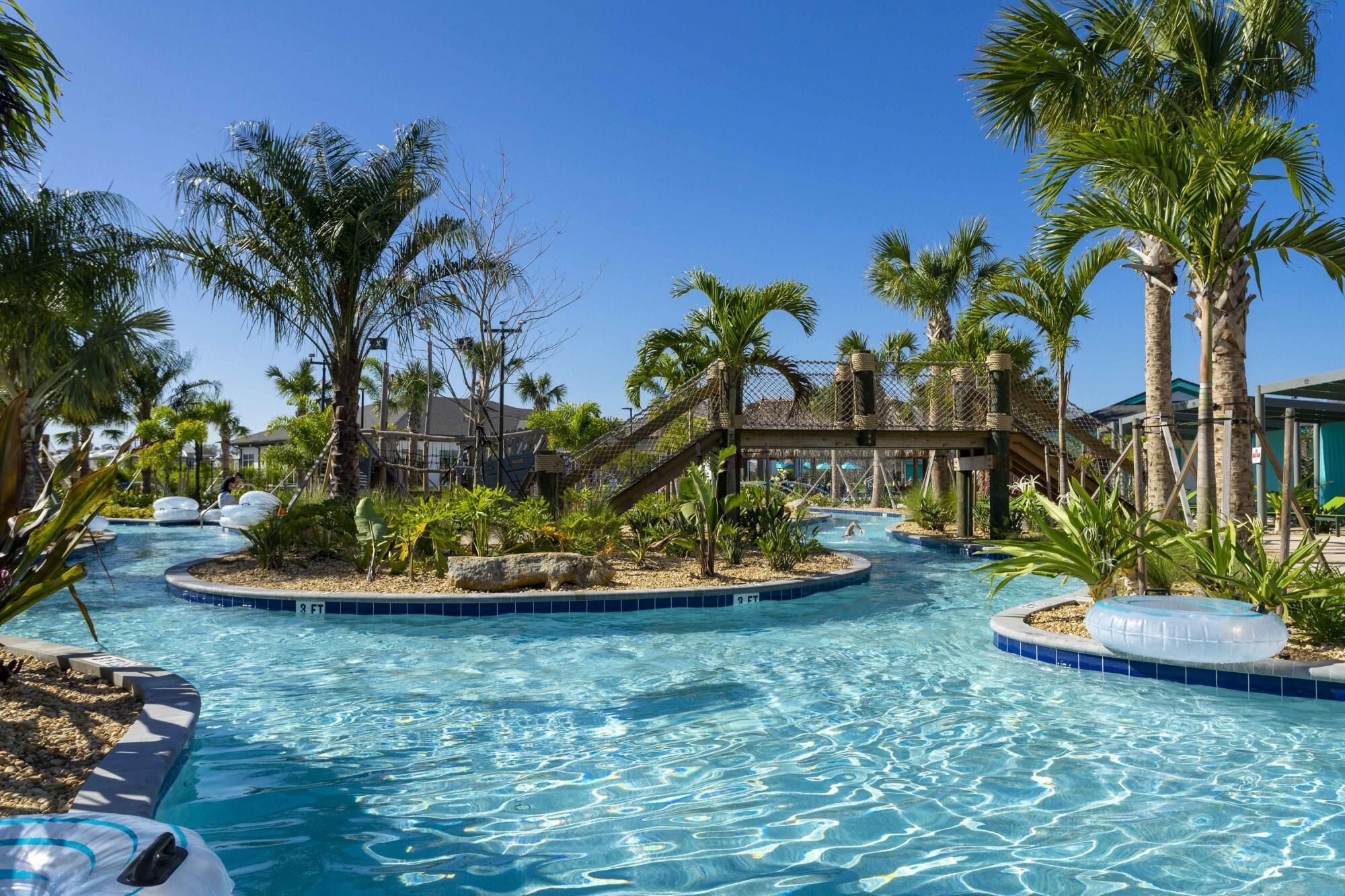 Un área de piscina con palmeras, un puente colgante y un pequeño edificio al fondo.