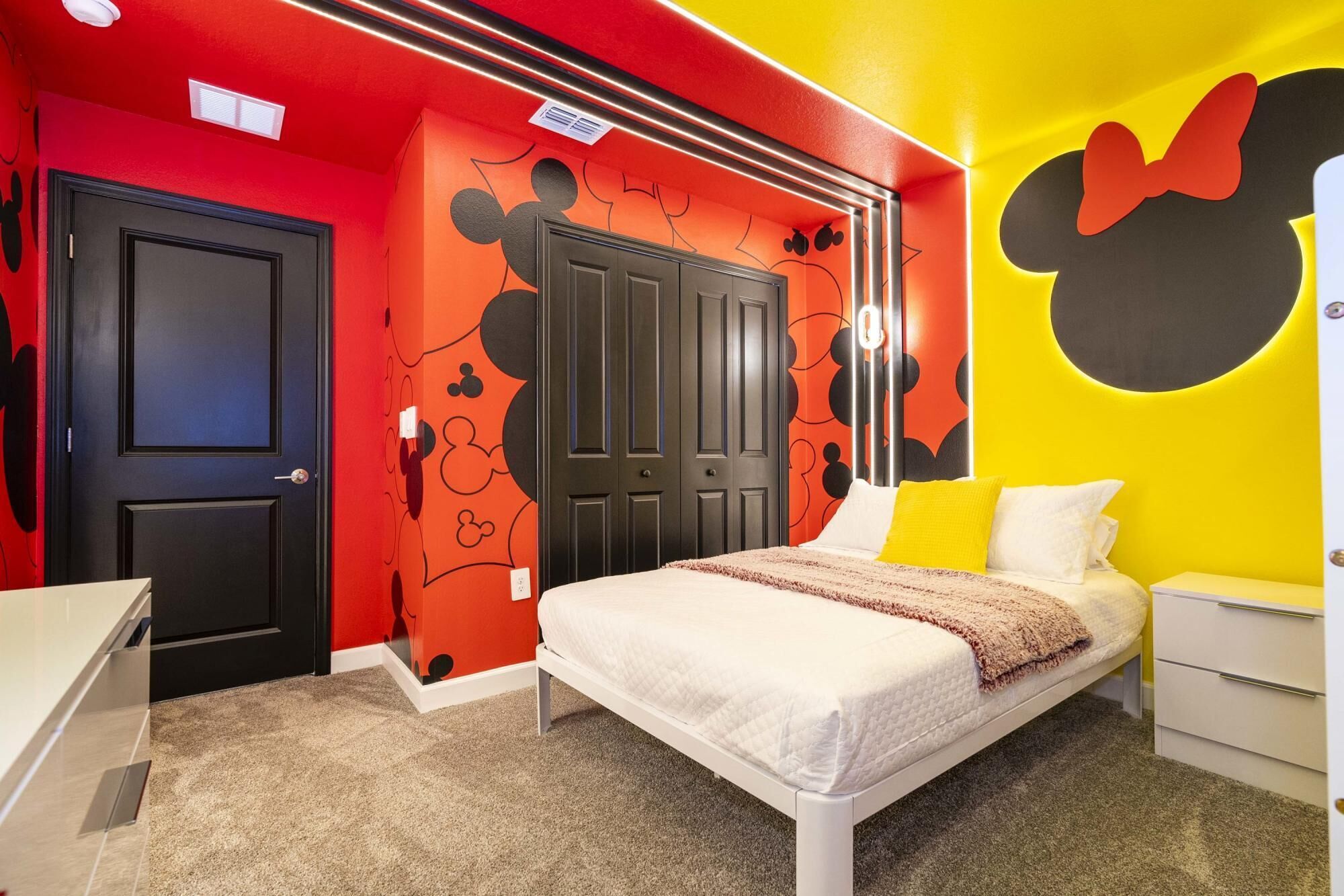 A bedroom with a bed, a desk, and a wardrobe. The walls are decorated with Mickey Mouse designs.