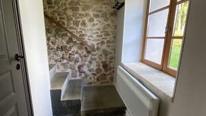 Un couloir étroit avec un mur en pierre, un escalier en bois, un radiateur et une fenêtre donnant sur la verdure.