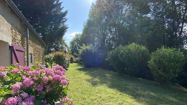 Un jardin avec un bâtiment en pierre, un ciel dégagé et un soleil qui filtre à travers les arbres.