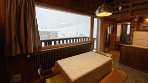 Interior acogedor de una cabaña con una mesa de comedor de madera, un banco, una ventana que ofrece vistas de un paisaje nevado y una zona de cocina con un refrigerador.