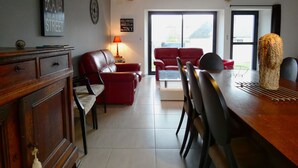 A living room with a red sofa, a wooden dining table, and a lamp. There is a wall clock and a sign with city names. - Charming Contemporary Farm Gîtes in Scenic Beauce Village, Private Garden & Parking (Moutiers)