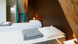 A bedroom with two beds, a wooden ceiling beam, a wall-mounted lamp, and a bathroom visible through a glass door. - Charming Contemporary Farm Gîtes in Scenic Beauce Village, Private Garden & Parking (Moutiers)